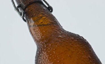 Condensate drops flowing bottle with cold beer in super slow motion close up. Clear condensate drops flowing down glass bottle with fresh cold beer in super slow motion close up. Brown glassware for refreshing drinks covered water droplets on white background. Freshness concept