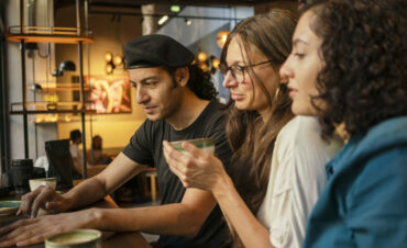 Three adults sit at a cafe table, one man working on a laptop and two women engaged in a conversation, with coffee cups in front of them, Berlin, Germany