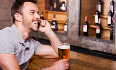 Waiting for you, pal! Side view of cheerful young man holding glass with beer and talking on the mobile phone while sitting at the bar counter Waiting for you, pal! Side view of cheerful young man holding glass with beer and talking on the mobile phone while sitting at the bar counter